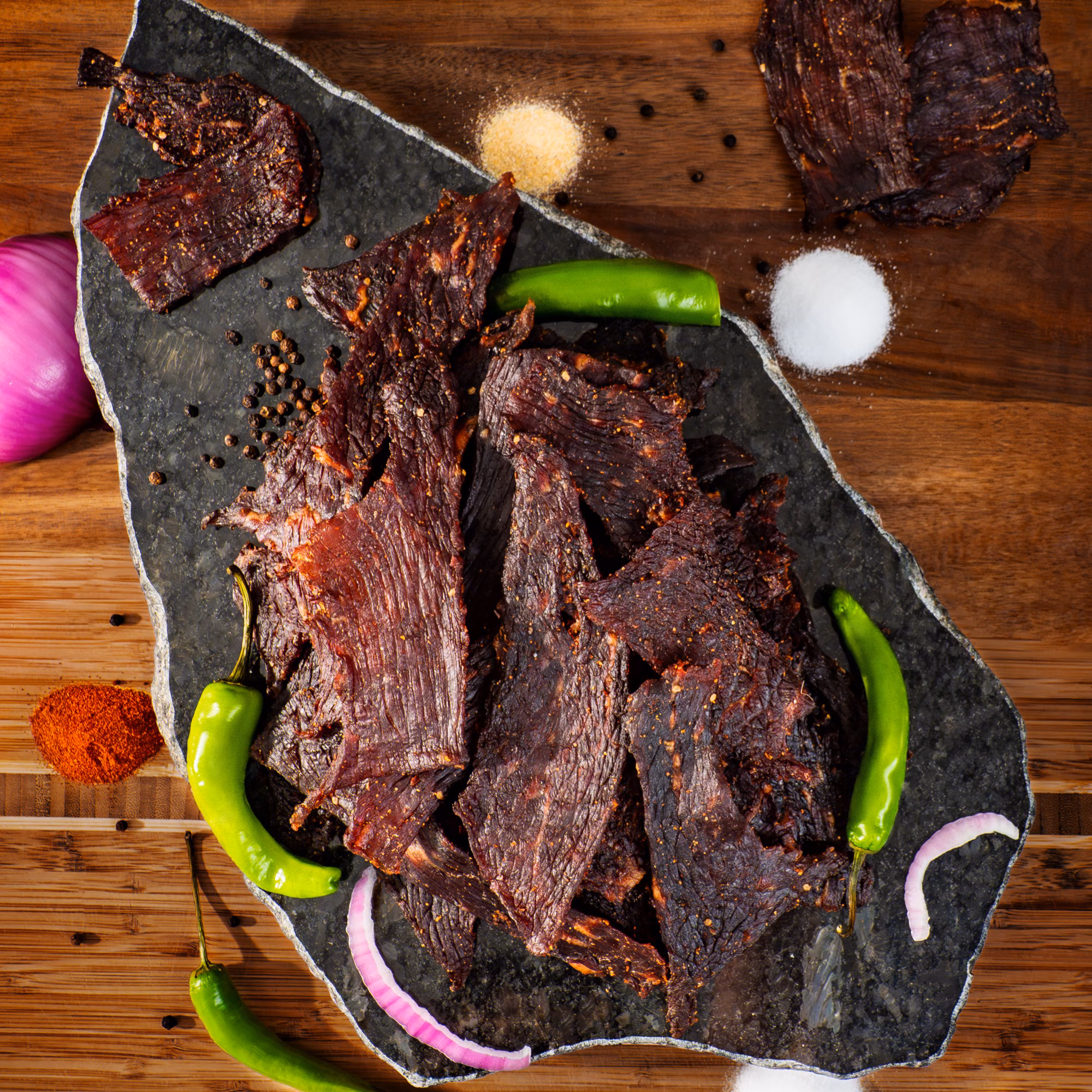 Matt-Hat Jerky pieces arranged on a stone board with green peppers, onion, salt, pepper, and spices.