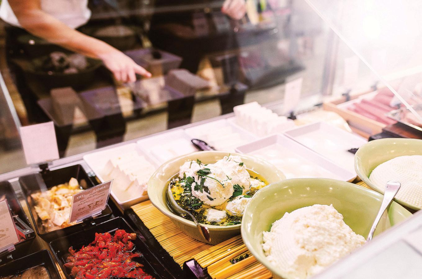 Display of various cheeses and spreads in bowls and trays behind glass, with a person’s arm pointing at items.