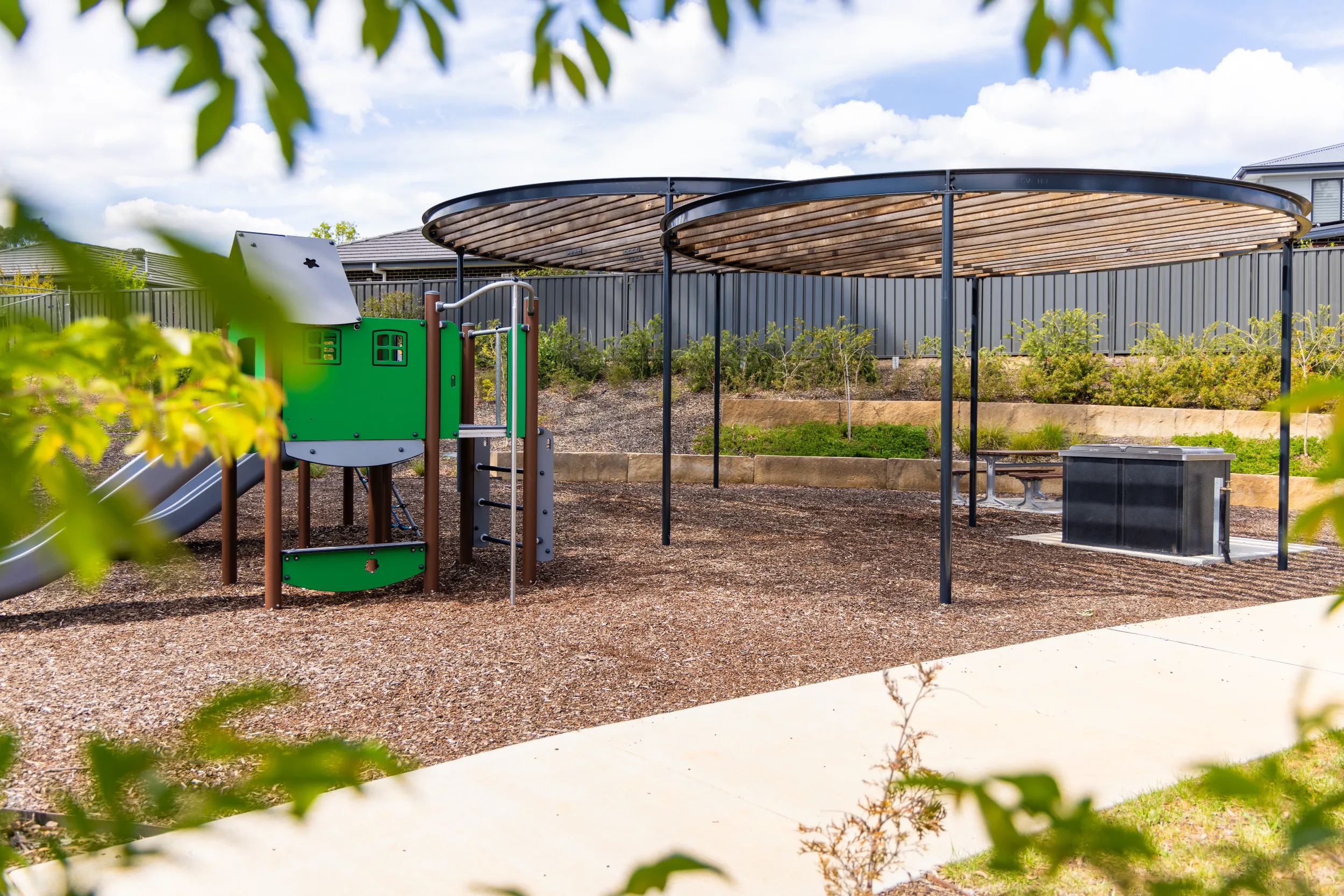 Playground area with a green play structure and a large circular wooden shade canopy over picnic tables and a barbecue grill.