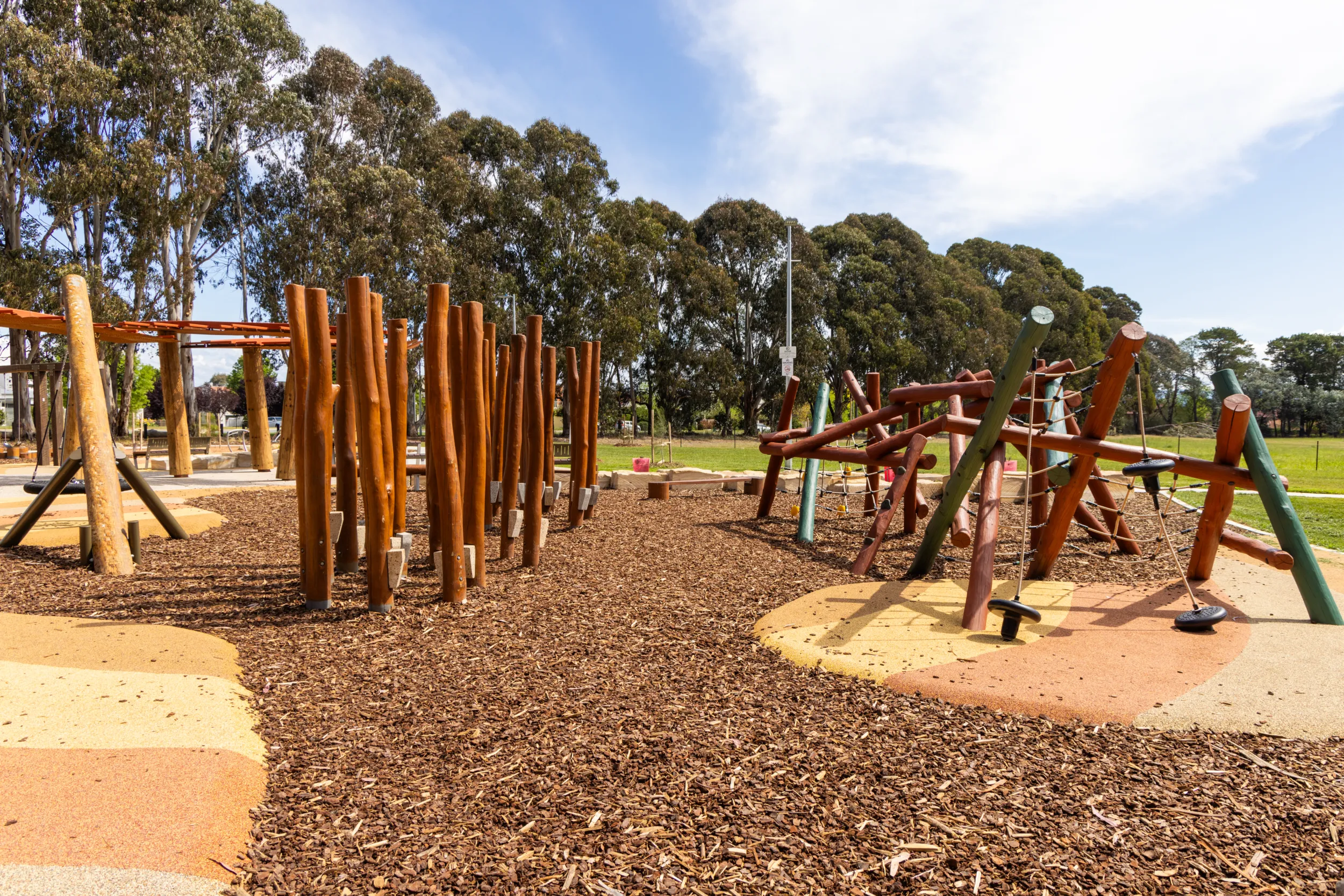 Outdoor playground with wooden climbing posts, rope structures, and mulch ground covering under a partly cloudy sky.