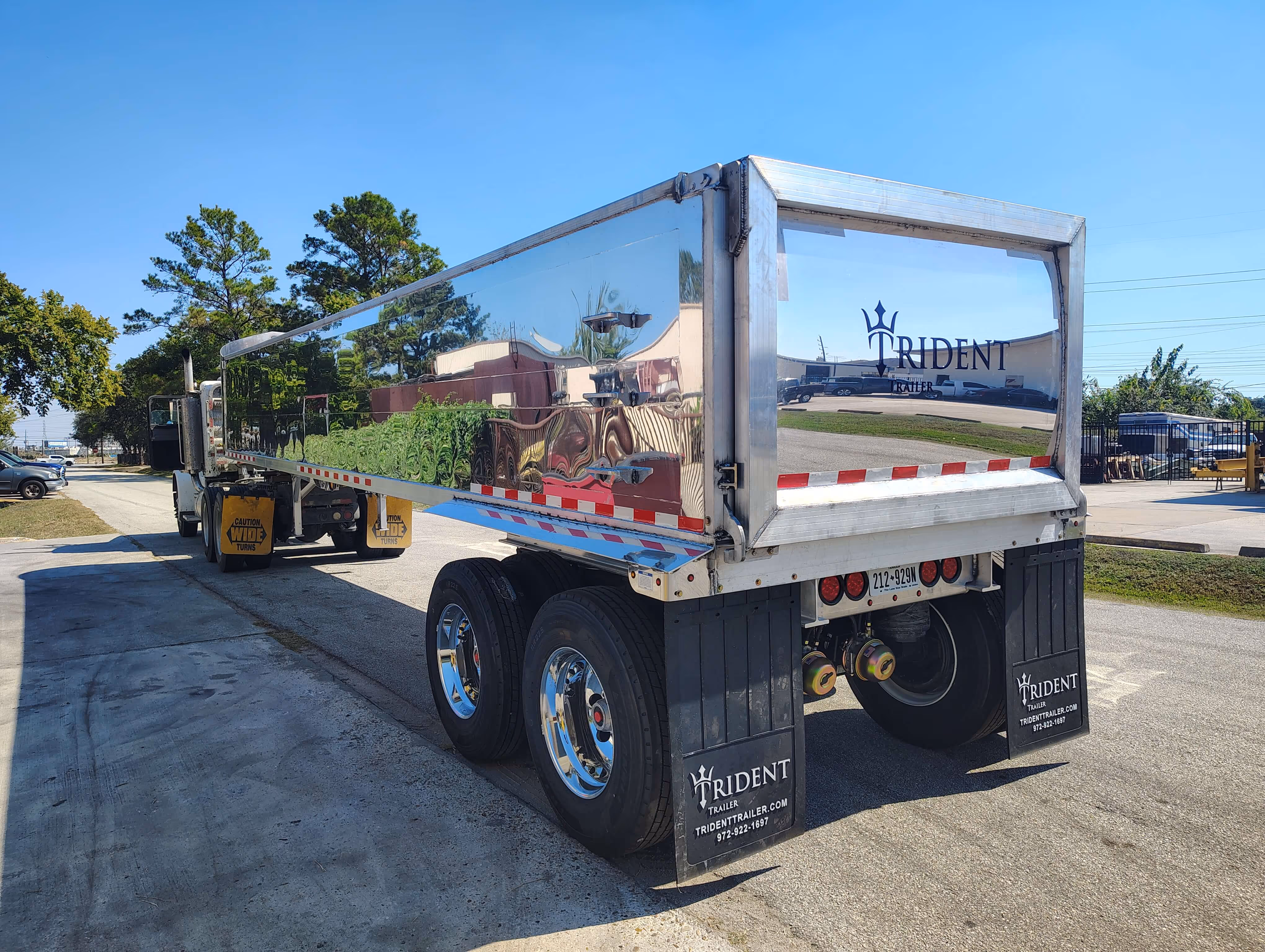 Rear side view of a shiny metallic Trident trailer attached to a semi truck parked on a street on a clear day.
