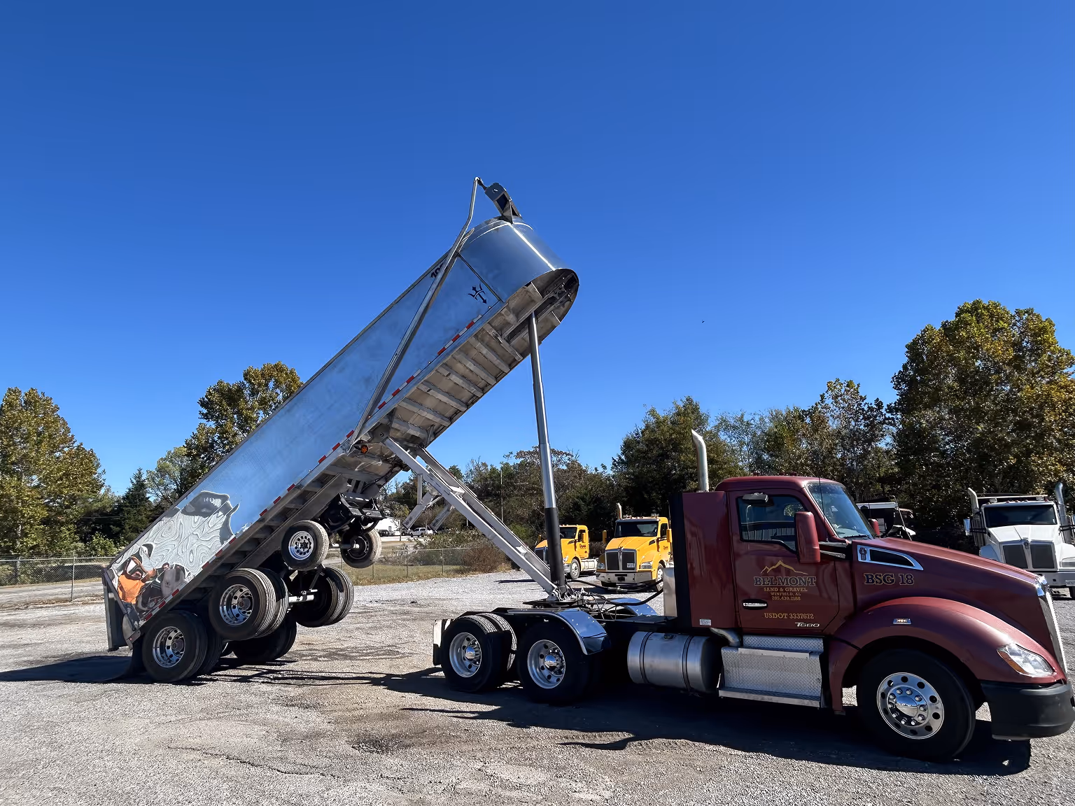 Maroon Belmont Sand & Gravel semi-truck with raised silver dump trailer in a parking lot under clear blue sky.