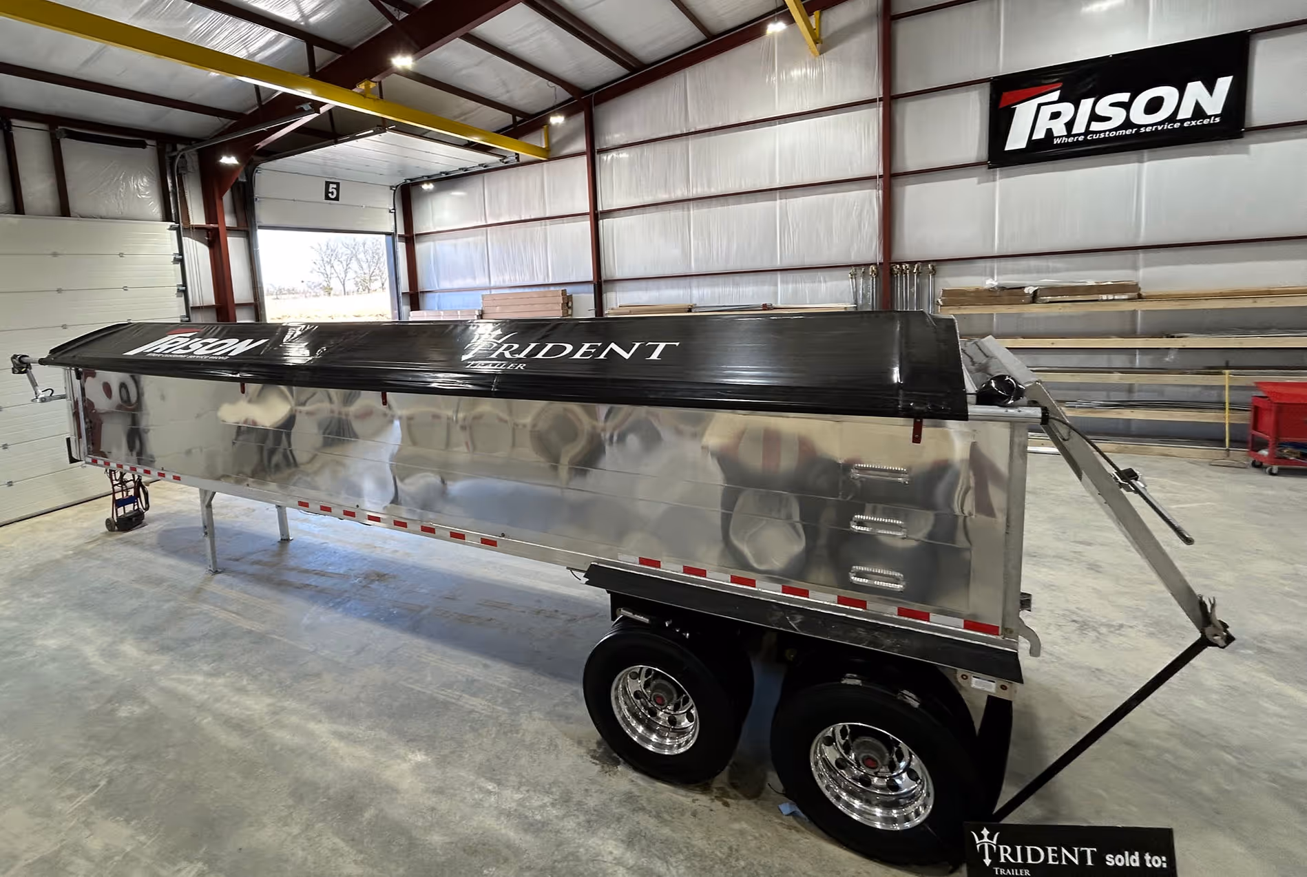 Polished metal trailer with black Trident cover inside a warehouse with Trison signage on the wall.