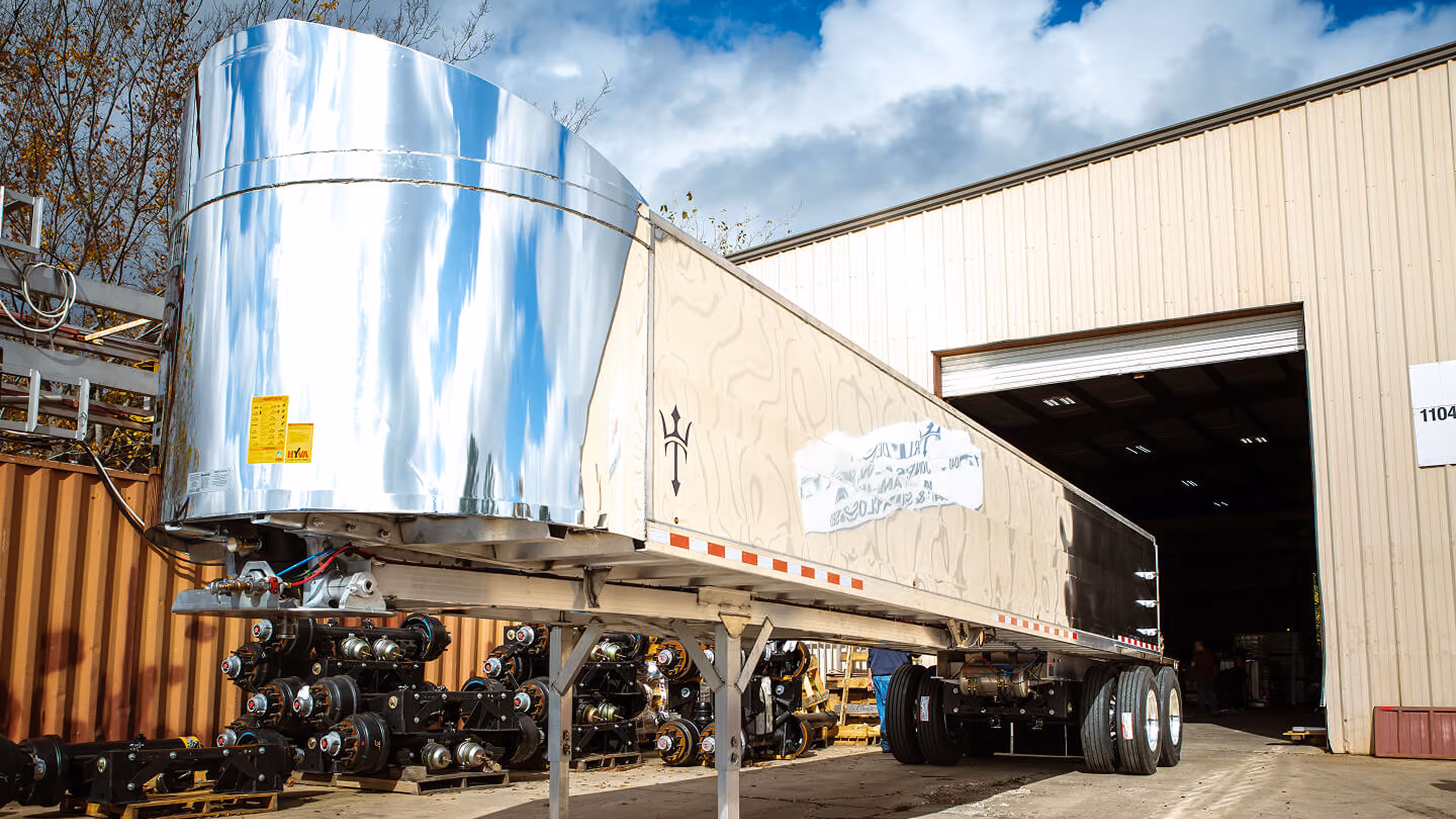 Shiny silver semi-trailer parked outside a warehouse with stacked axles in the background.