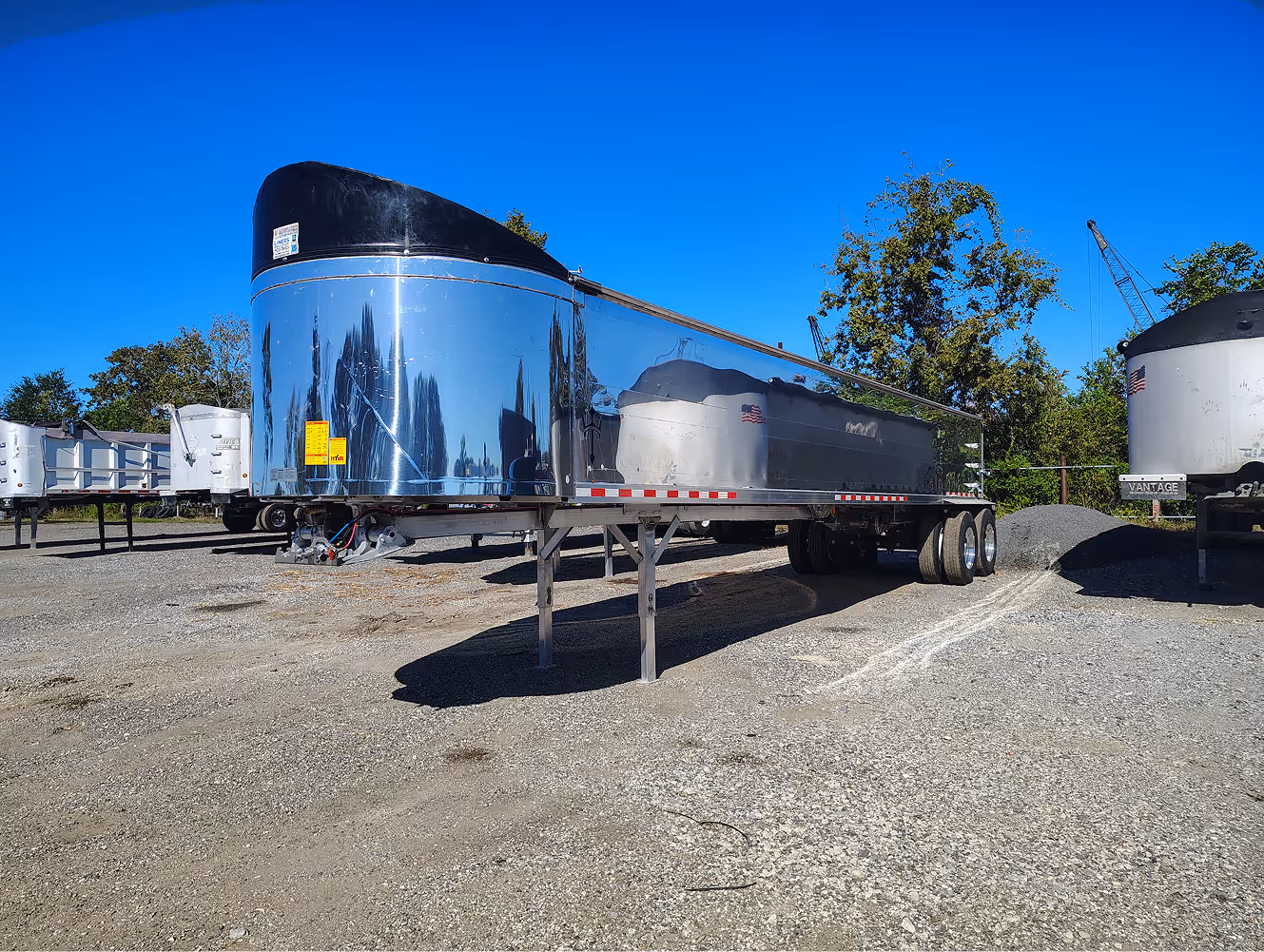 Shiny silver semi-trailer parked on a gravel lot under a clear blue sky.