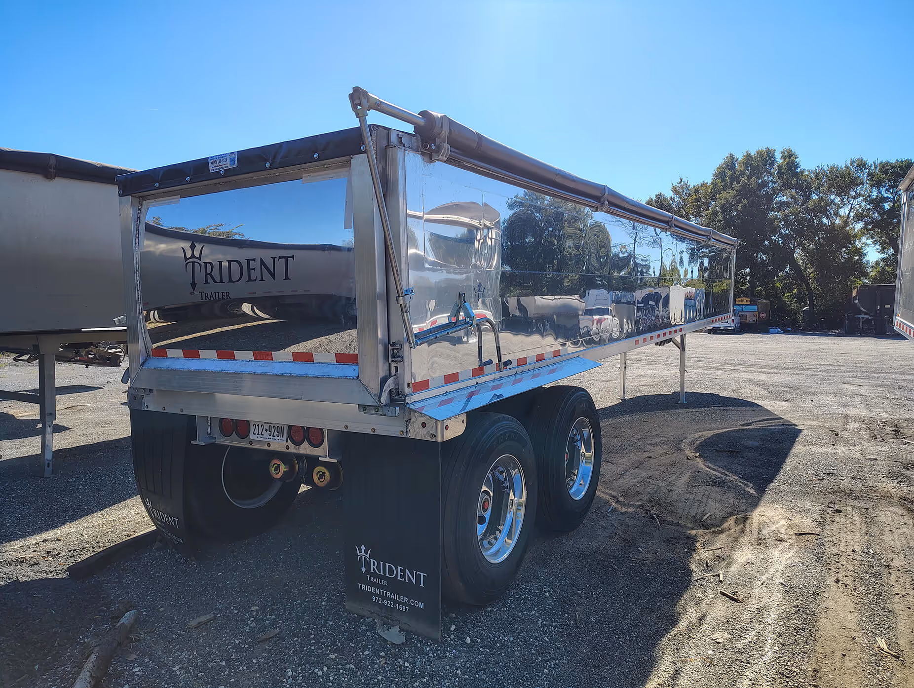 Shiny silver trailer with dual rear wheels parked on gravel under clear blue sky.
