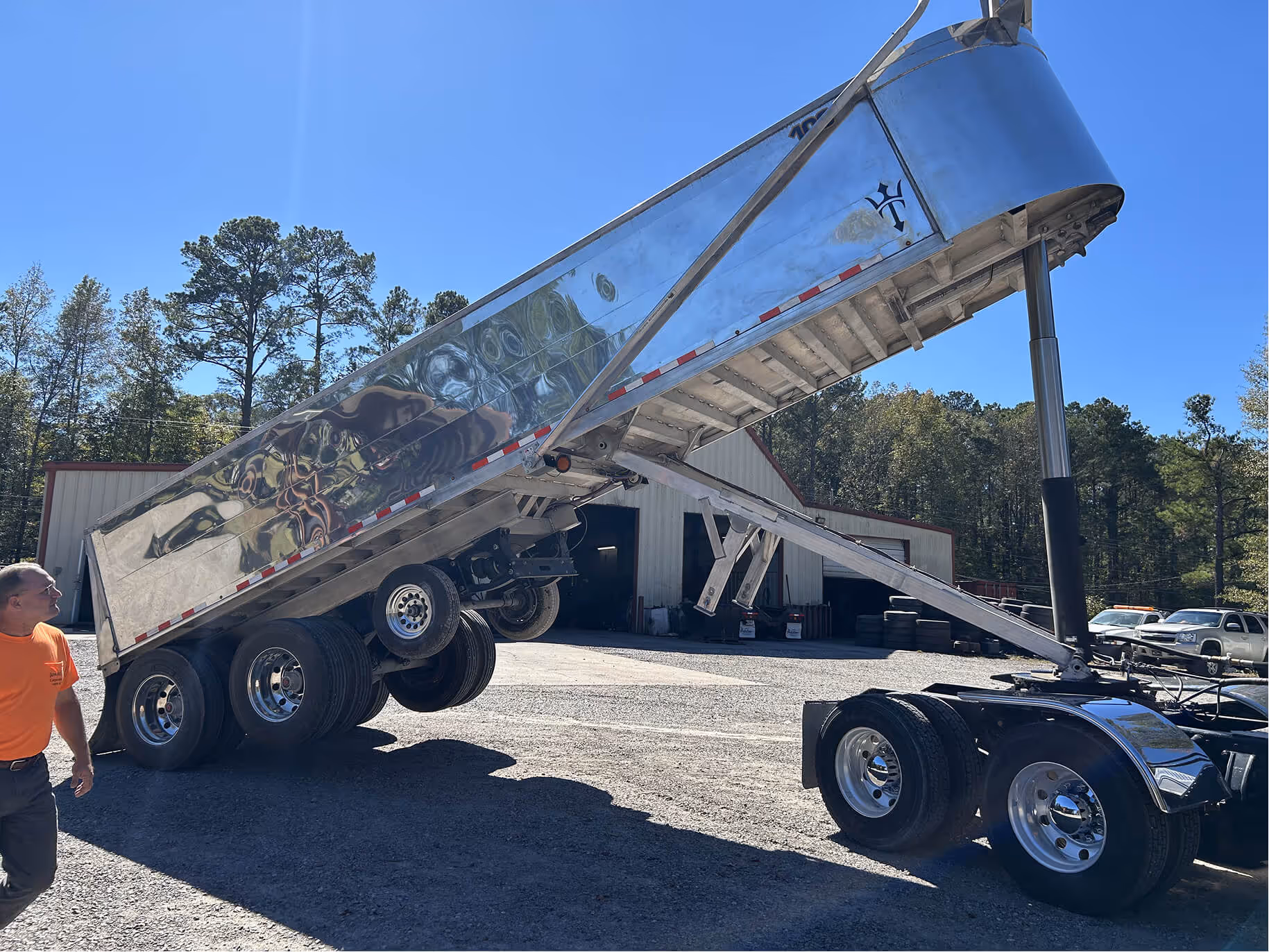 A silver dump truck trailer raised at an angle with a man in an orange shirt walking nearby in a gravel lot with buildings and trees in the background.