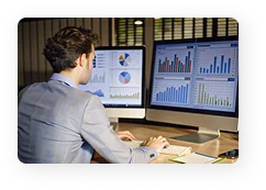 Man in a suit working at a desk with two monitors displaying various business charts and graphs.