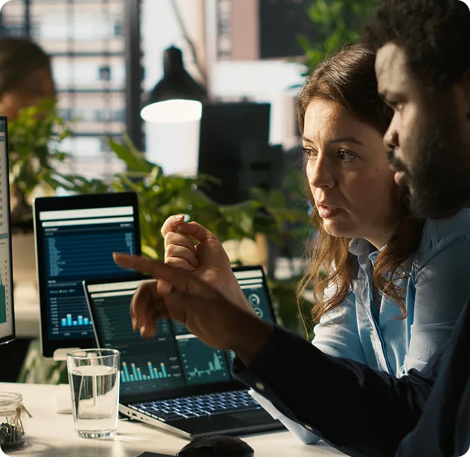 Two colleagues closely examining data charts on multiple laptop screens in an office setting.