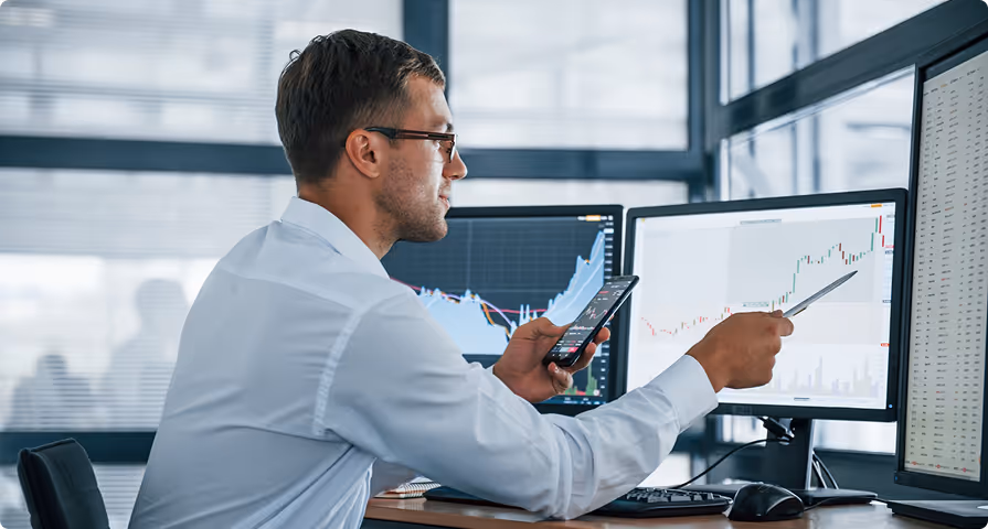 Man in glasses pointing at a stock chart on a computer monitor while holding a smartphone, surrounded by multiple monitors showing financial data.