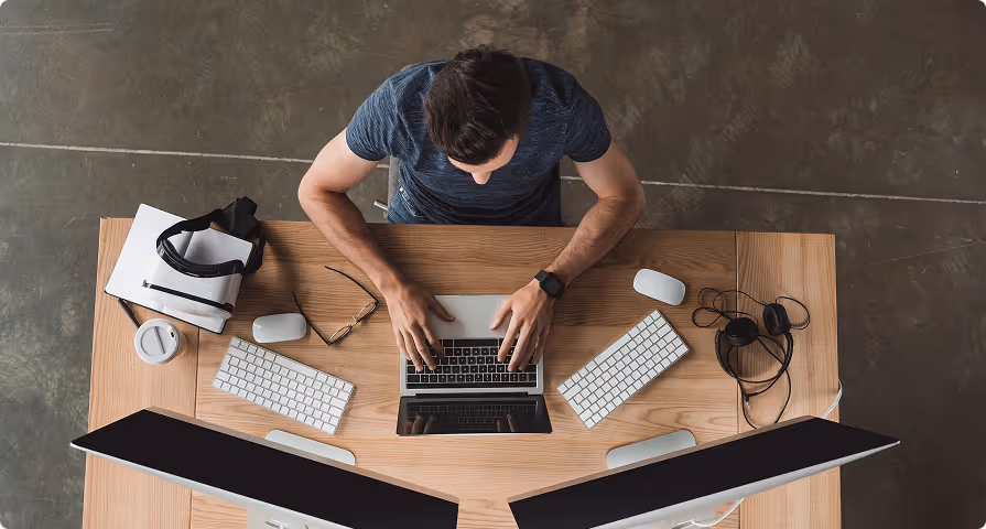 Top view of a person typing on a laptop at a wooden desk with two large monitors, keyboards, mouse devices, headphones, and glasses nearby.