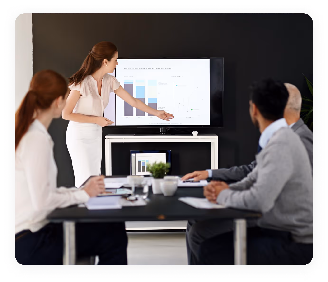 A woman presenting sales strategy charts on a screen to three colleagues seated at a conference table.