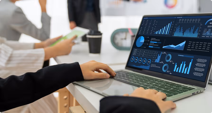 Person working on a laptop displaying various blue data charts and graphs at a white desk with two people in the background.
