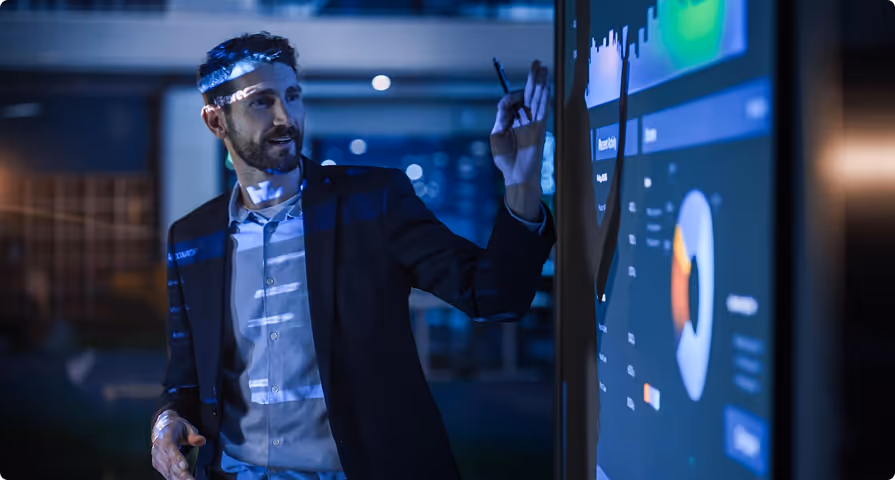 Man in business attire writing on a large digital screen displaying charts and graphs in a dark office setting.