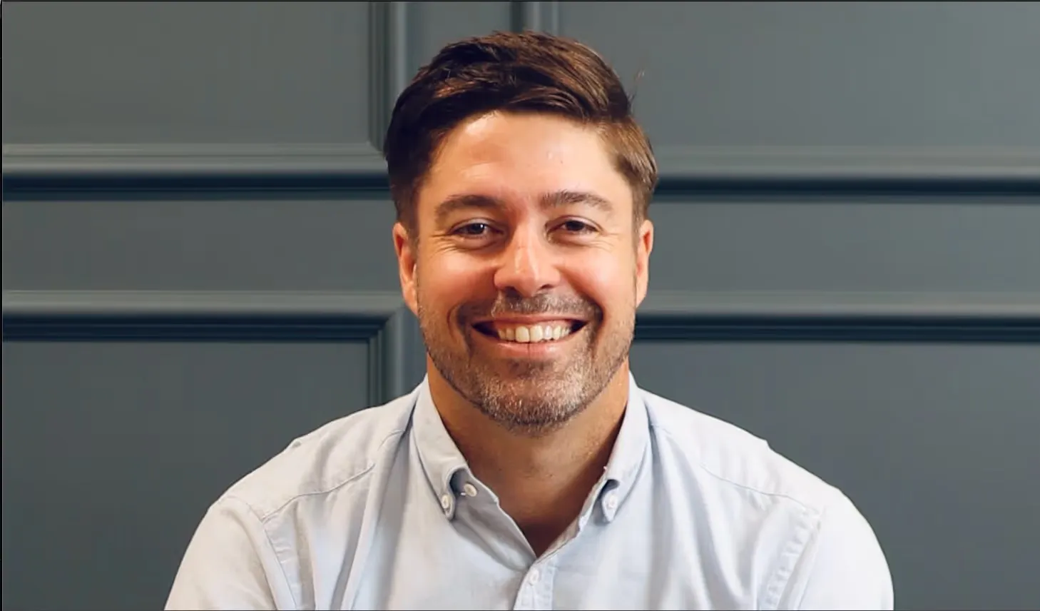 Smiling man with neatly combed hair and a light beard wearing a light blue button-up shirt against a gray paneled wall.