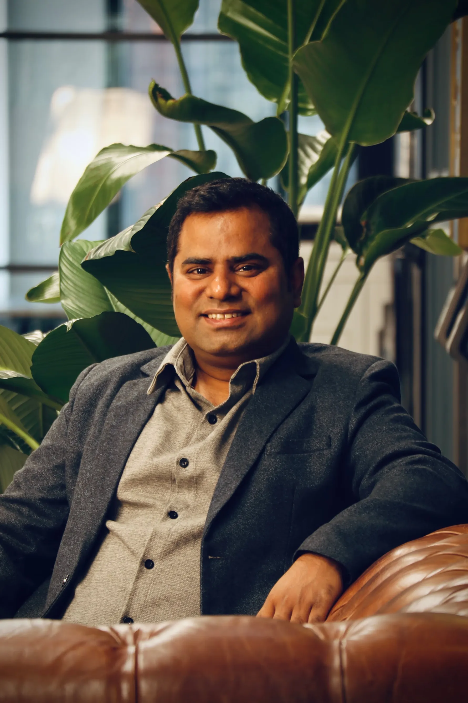 Smiling man wearing a gray blazer and shirt sitting on a brown leather sofa with large green plants in the background.