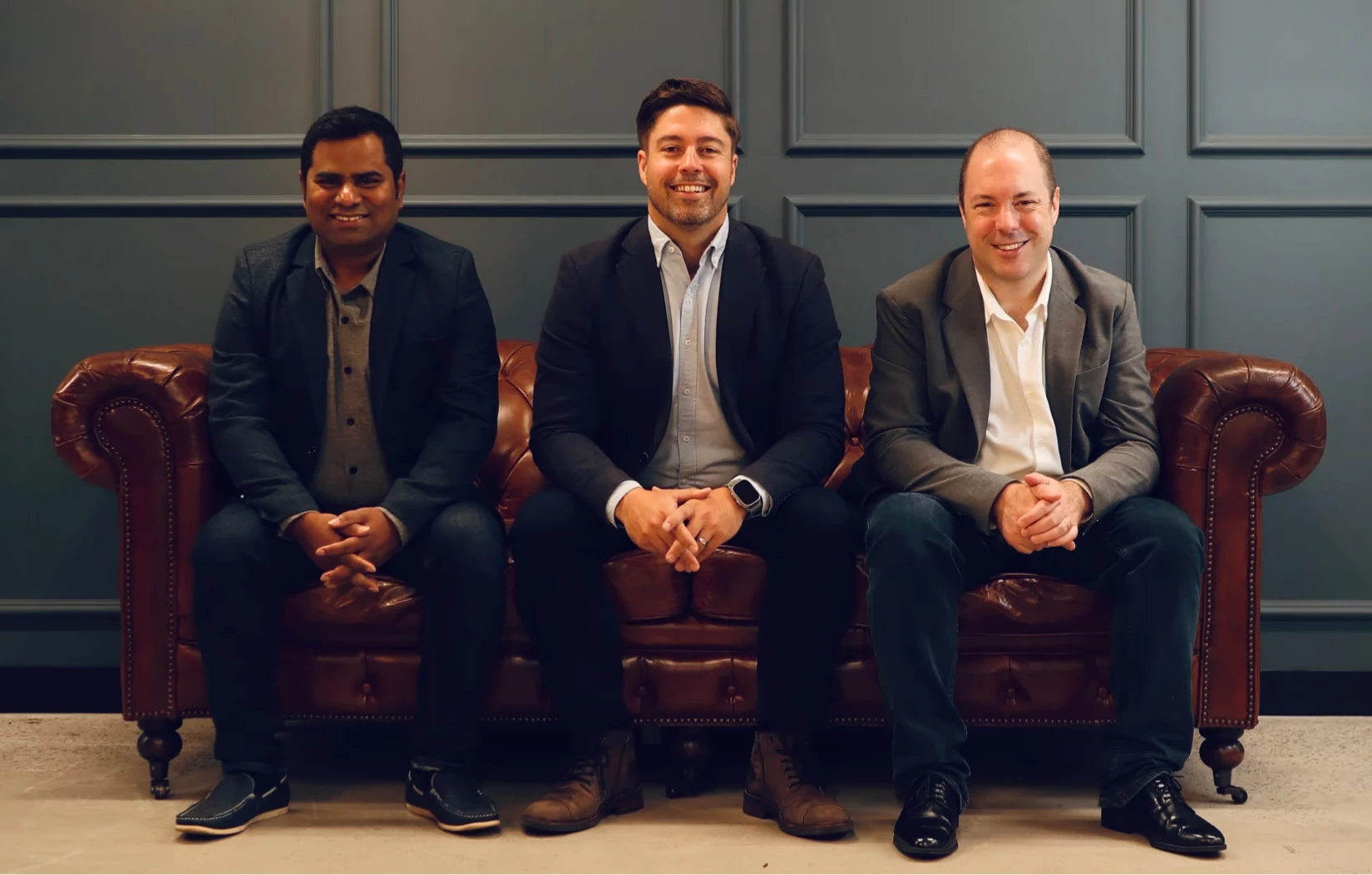 Three men sitting on a brown leather sofa against a gray paneled wall, all smiling and wearing blazers.