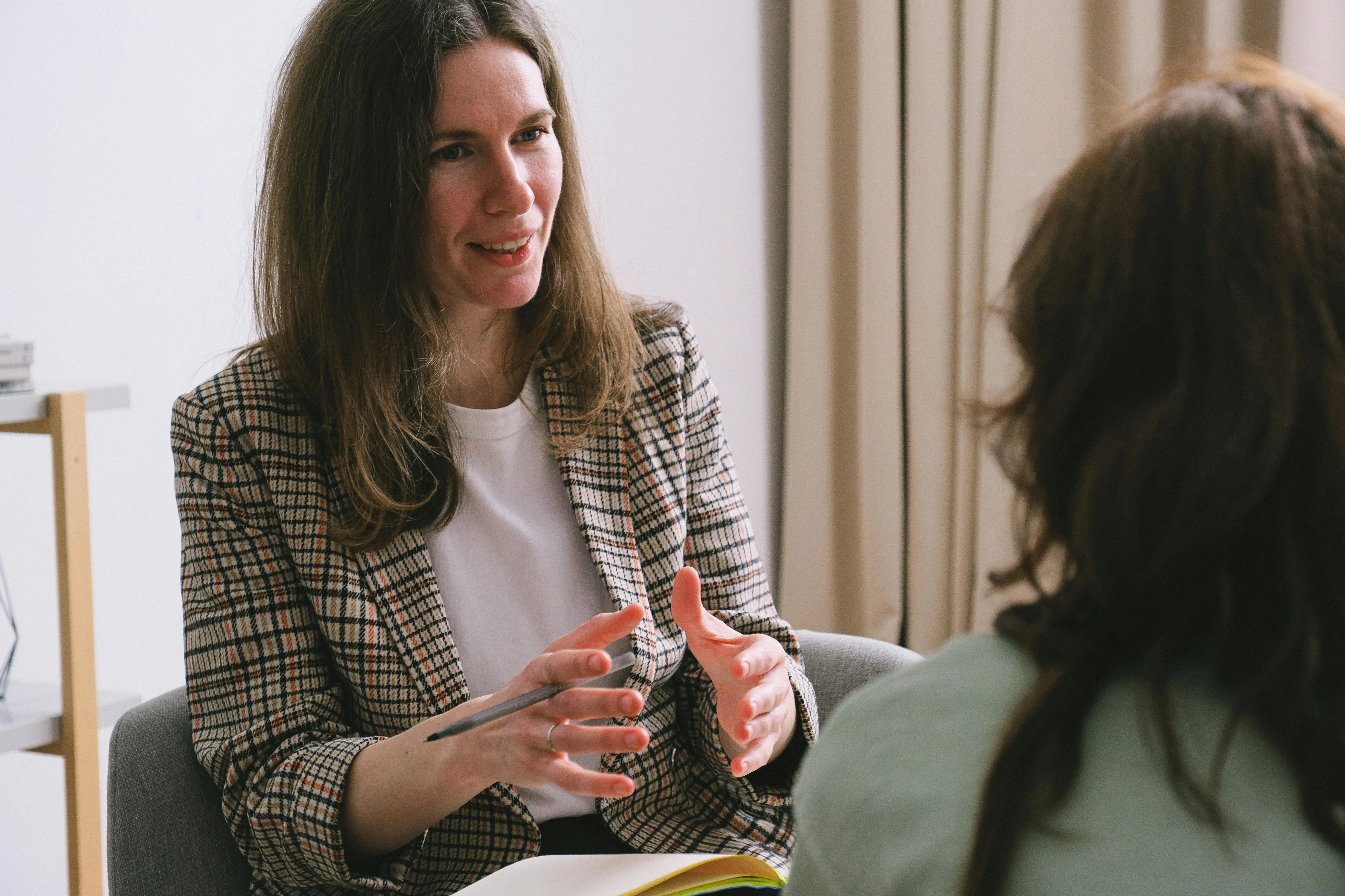 Woman in a checkered blazer gestures while talking to another person in a light-filled room.