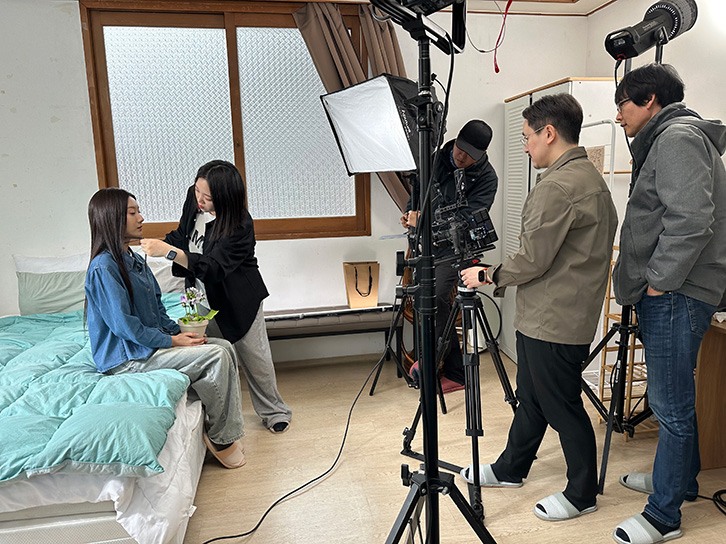 A woman sitting on a bed holding a potted plant while another woman adjusts her collar, with three men operating camera and lighting equipment in a small room.