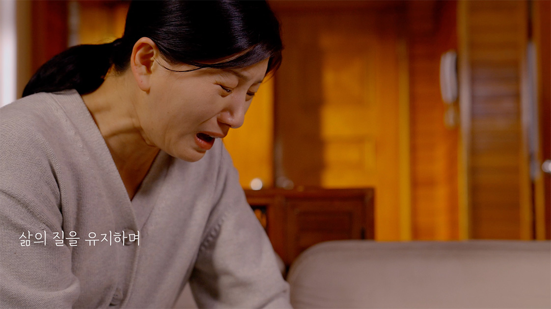 Woman in gray sweater crying indoors with Korean text on the left side.