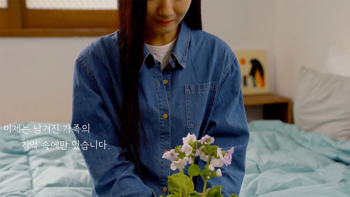 A woman in a blue denim shirt sitting on a bed, gently holding a pot of pink and white flowers.