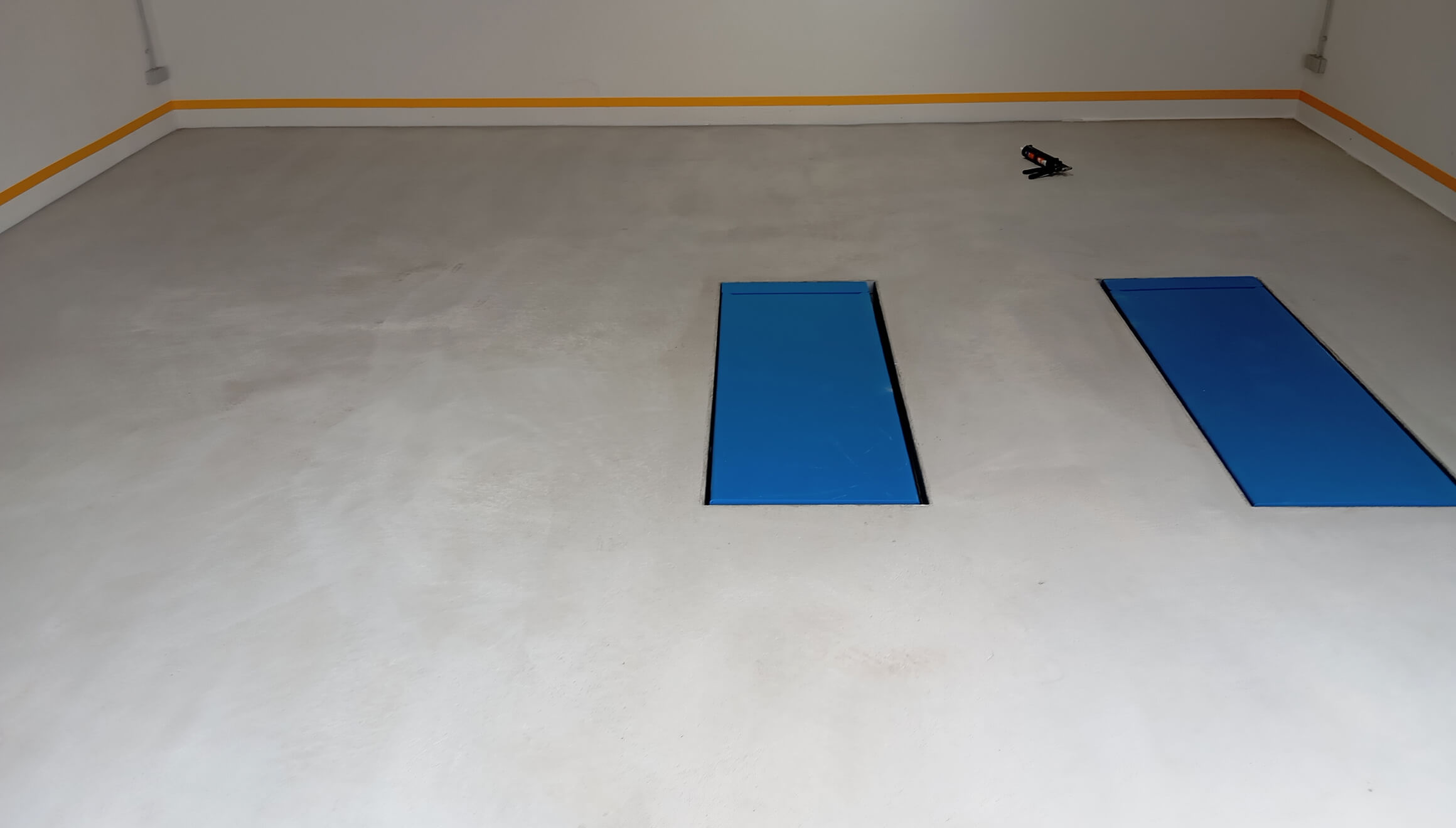 Leerer Raum mit grauem Boden und zwei blauen Bodenklappen, orangefarbene Streifen an den Wänden.