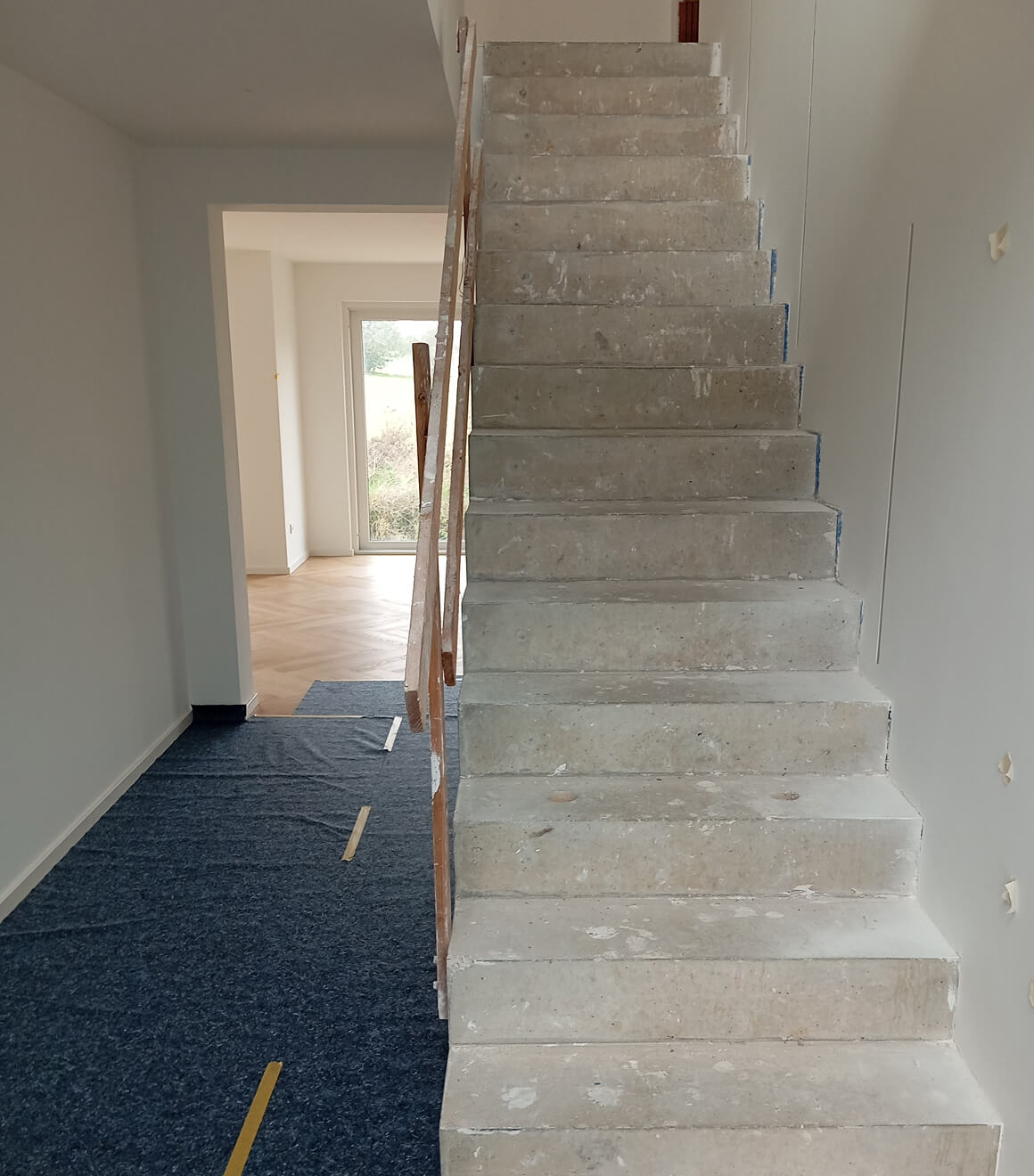 Unfertige Betontreppe in einem Innenraum mit einem blauen Schutzvlies auf dem Boden und einem Handlauf aus Holz.