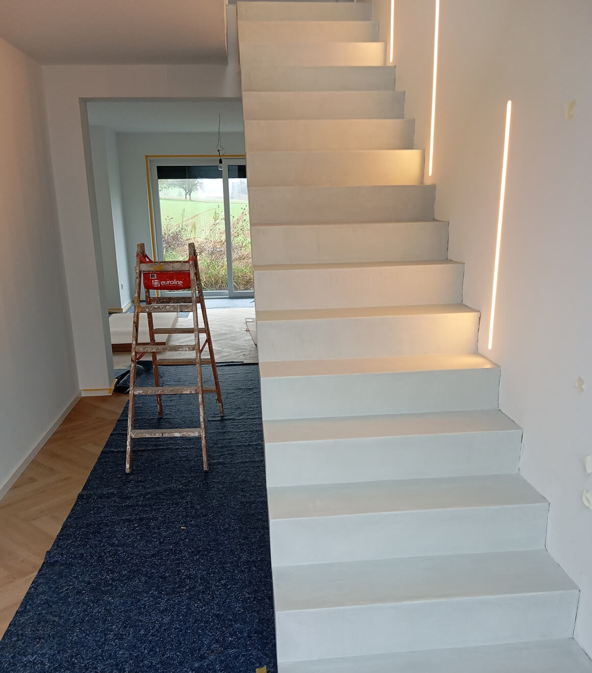 Innentreppen mit heller Oberfläche und integrierten LED-Lichtstreifen an der Wand, daneben eine gebrauchte Holzleiter auf einem dunkelblauen Teppich in einem hellen Raum mit großen Fenstern.