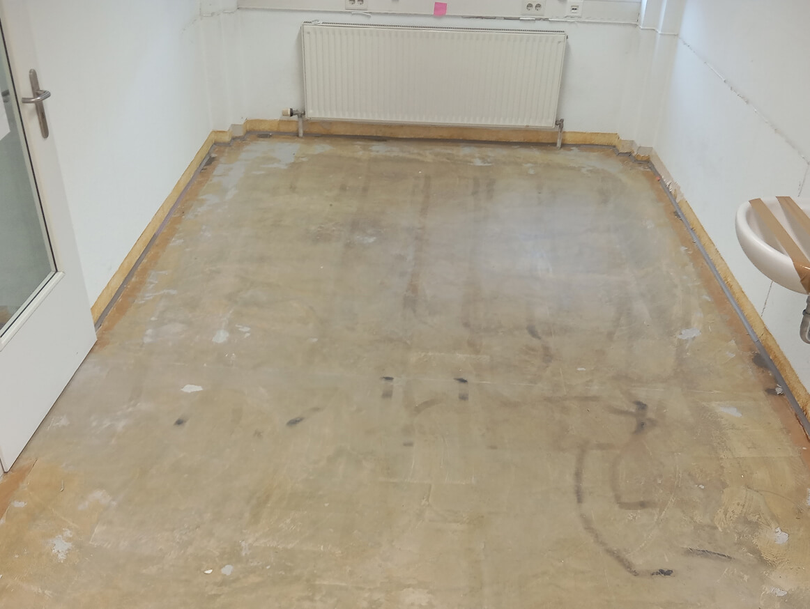 Leerer Raum mit kahlem Betonboden, weißen Wänden, einem Heizkörper an der Rückwand und einem weißen Waschbecken rechts.