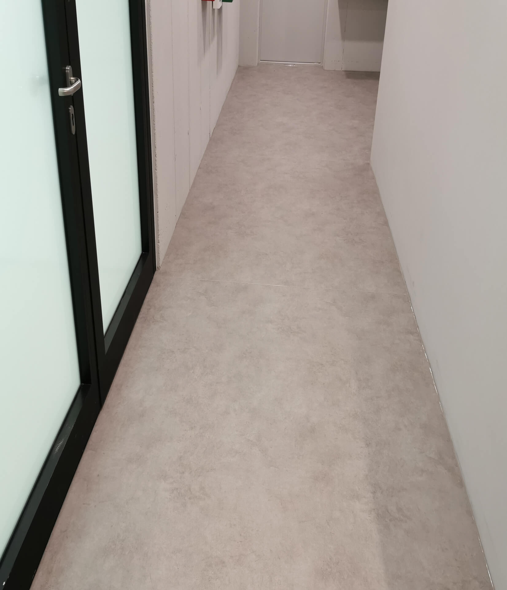 Schmaler Flur mit hellgraßem Bodenbelag, weißen Wänden und einer Tür mit Glas und schwarzem Rahmen links.