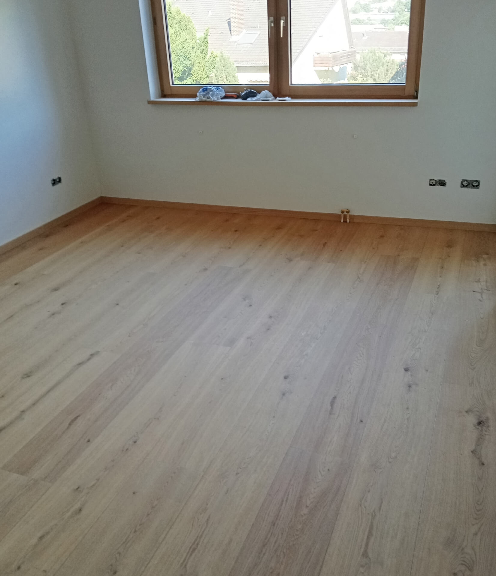 Leerer Raum mit hellem Holzboden, weißen Wänden und einem Fenster mit Blick auf Bäume und Häuser.