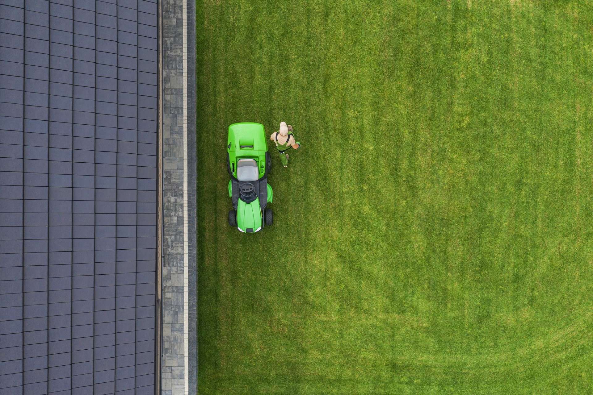Overhead view of a person using a green ride-on mower to cut a well-manicured lawn next to a house with a dark tiled roof. The freshly cut grass shows neat lines, emphasizing the precision and care in the lawn mowing process.