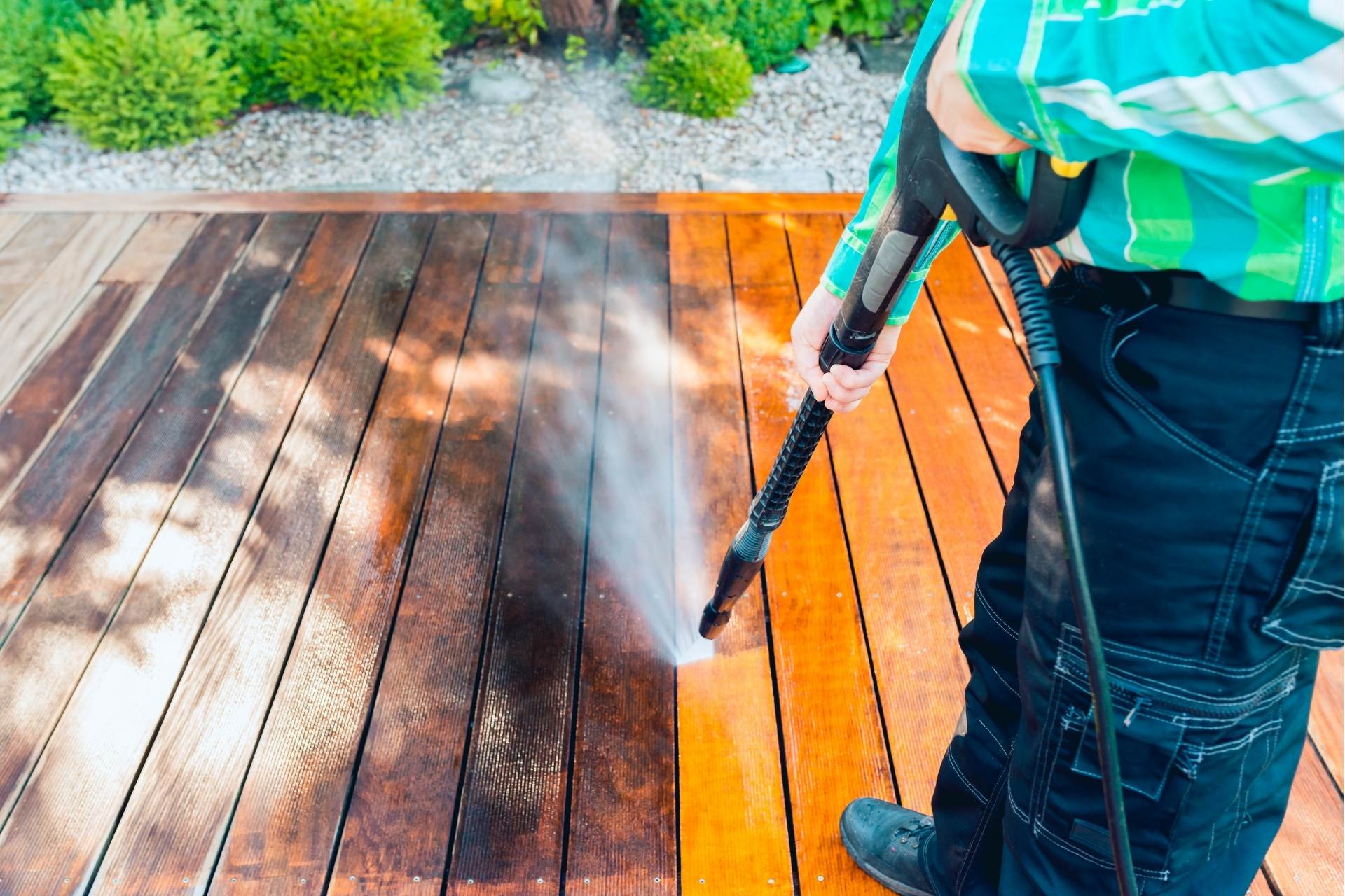A man using a high-pressure washer to clean a wooden terrace, with water spraying across the planks as dirt lifts away. Sunlight highlights the difference between the cleaned and uncleaned sections of the deck.