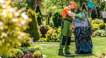 A person in work gloves and protective clothing is emptying the grass catcher of a lawn mower after mowing.