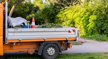 A person in work gloves and protective clothing is emptying the grass catcher of a lawn mower after mowing.
