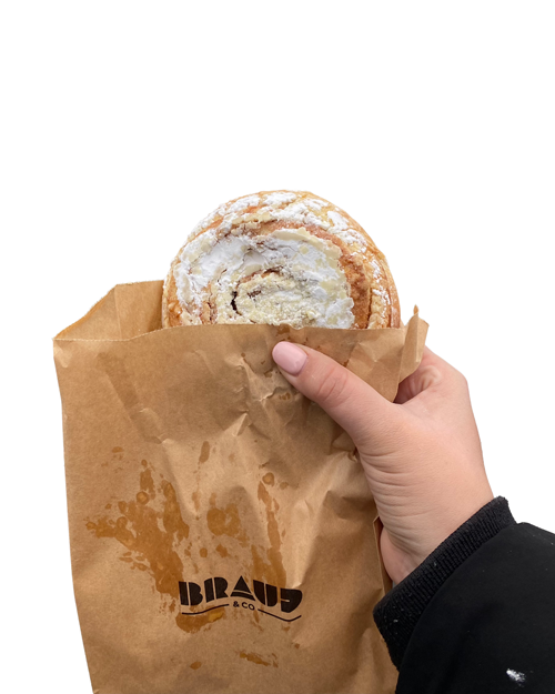 Hand holding a cinnamon roll wrapped in a brown paper bag with BRAUD & CO branding.