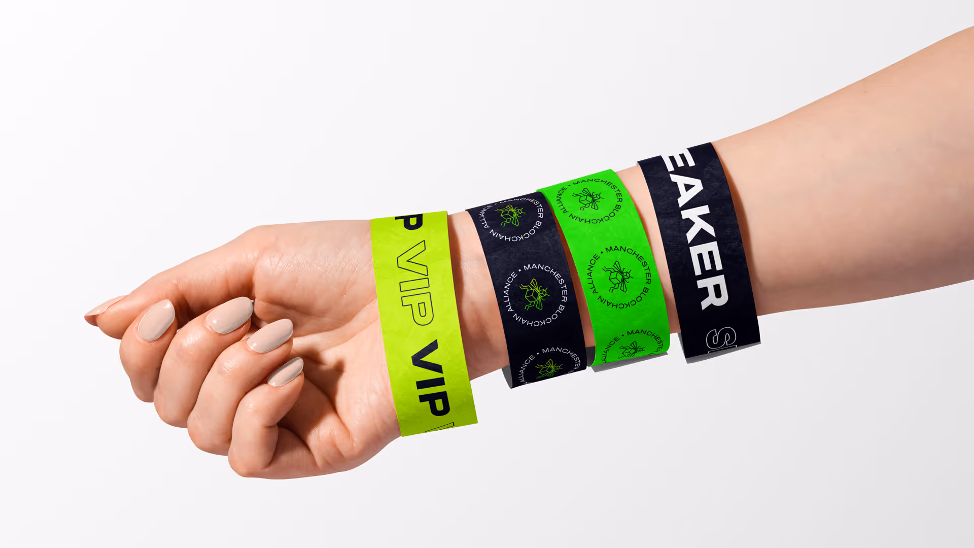 Left wrist wearing four event wristbands: a bright yellow-green VIP band, two bands with Manchester Blockchain Alliance logos in black and green, and a black speaker band.