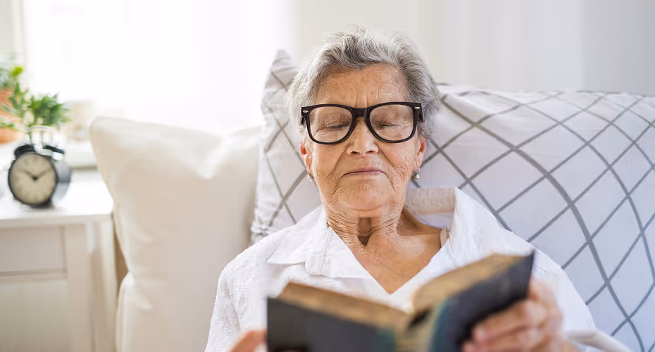 Senior souriante entrain de lire un livre sereinement dans son canapté grâce à son logement sécurisé