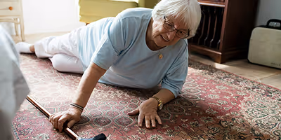 Une femme âgée, vêtue d'un tee-shirt bleu, est tombée sur un tapis dans un salon et tente de se relever en s'appuyant sur sa canne.