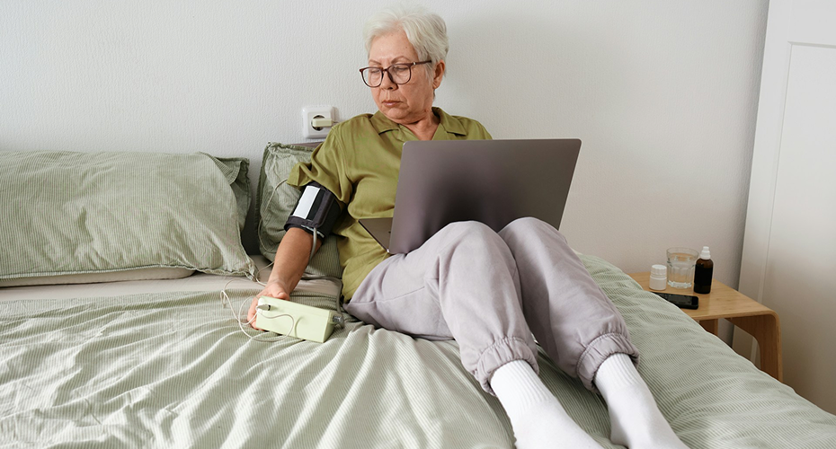 Femme senior à son domicile utilisant un tensiomètre connecté et un ordinateur pour son suivi de santé.