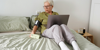 Femme senior à son domicile utilisant un tensiomètre connecté et un ordinateur pour son suivi de santé.