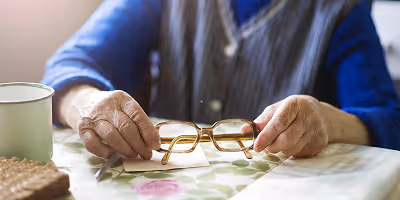 Personne âgée prenant sa paire de lunette dans les mains pour étudier les démarches administratives à effectuer pour obtenir des subventions pour l'aménagement de son domicile.
