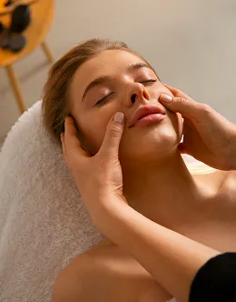 Woman receiving a gentle facial massage while lying on a white towel in a spa setting.
