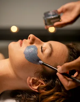Close-up of a woman lying down with a blue facial mask being applied to her cheek with a brush.