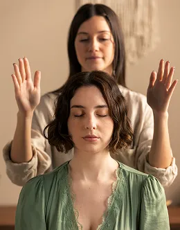 Woman with closed eyes sitting peacefully while another woman stands behind her with hands raised, in a calm indoor setting.