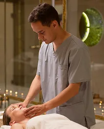 Therapist in gray scrubs giving a facial massage to a client lying down with eyes closed.