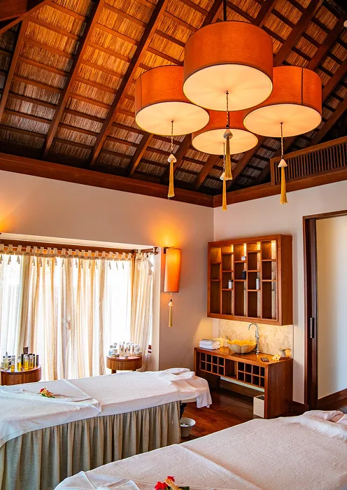 Spa room with two massage beds, wooden ceiling, warm lighting, and shelves with towels and spa products.