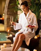 Woman in white bathrobe sitting by a pool holding a glass of champagne and a plate of strawberries, surrounded by lit candles and plants.