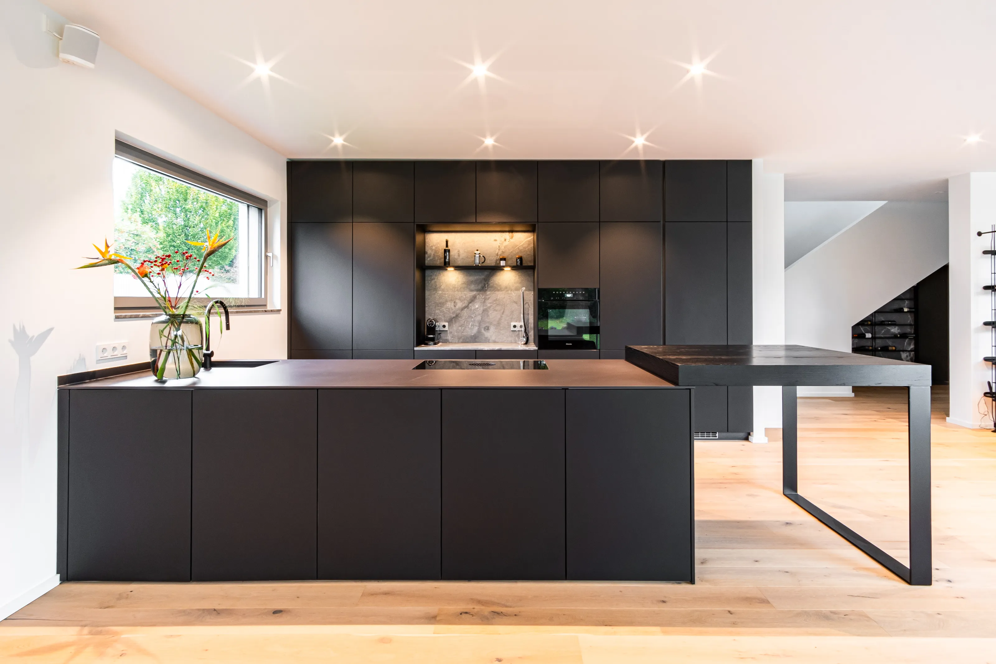 Minimalistische schwarze Küche mit Insel, eingebautem Herd, großem Fenster und Holzboden.