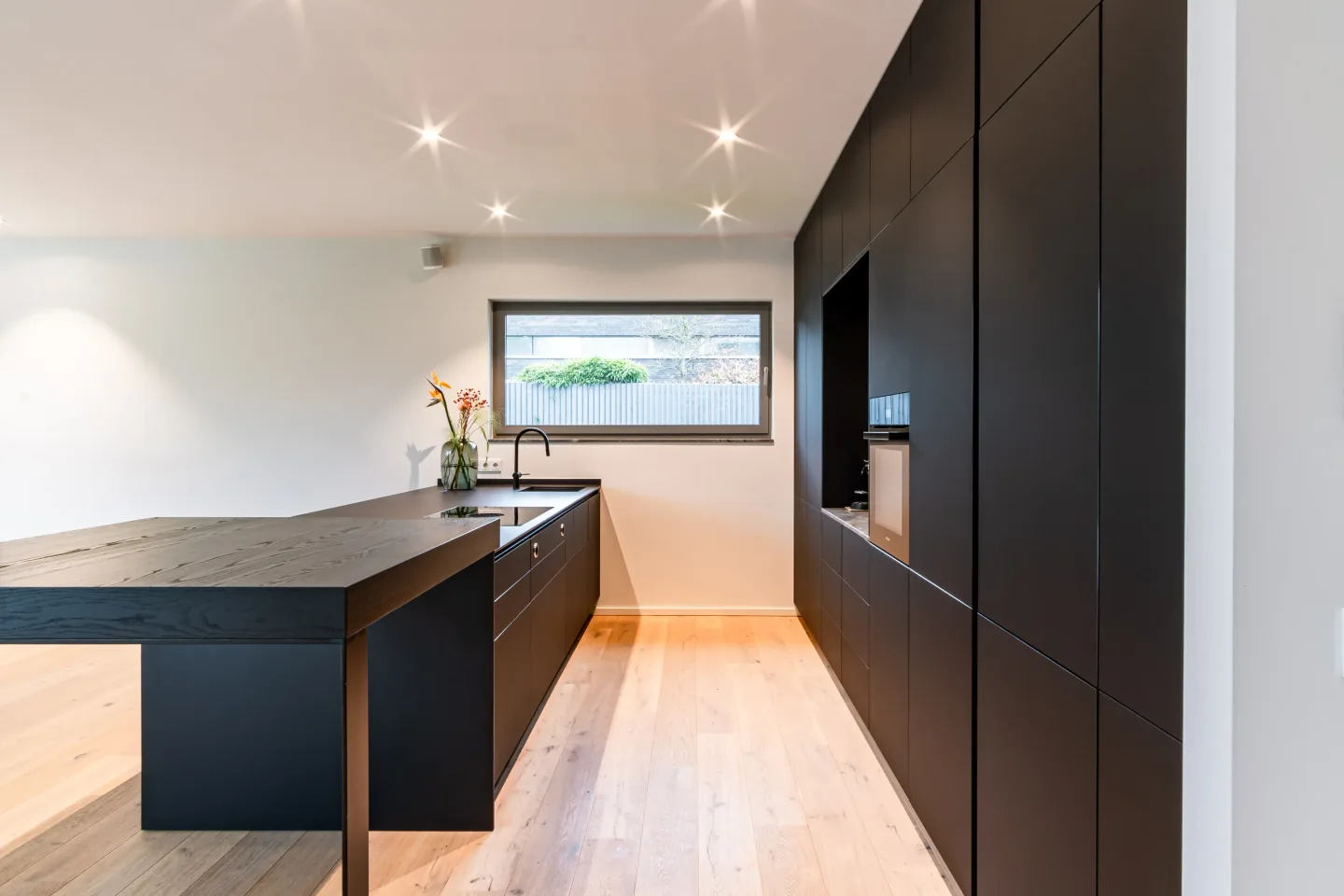 Schwarze minimalistische Küche mit Insel, Holzfußboden und Fenster mit Blick ins Freie.